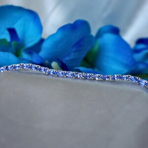 Op de afbeelding: Een delicate zilveren armband met een rij ovale blauwe edelstenen. De armband ligt op een witte stoffen achtergrond.