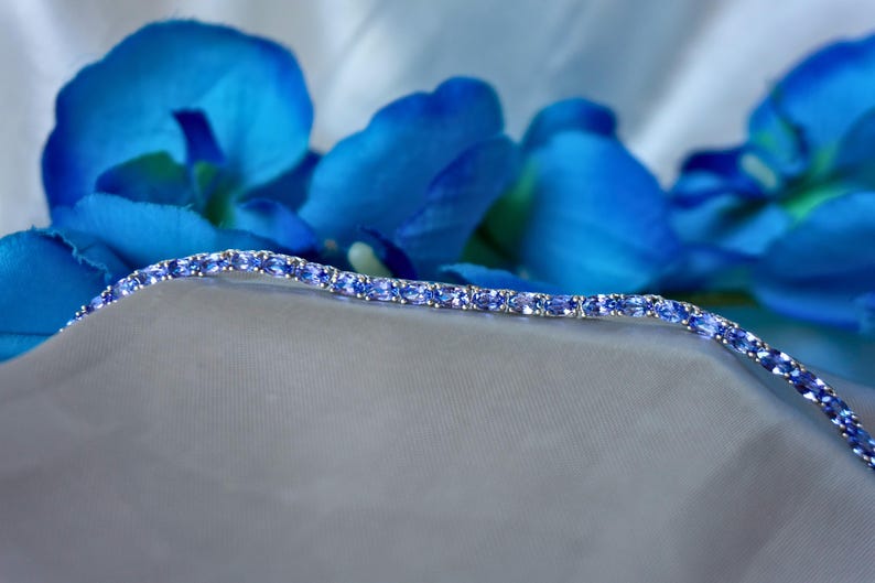 Op de afbeelding: Een delicate zilveren armband met een rij ovale blauwe edelstenen. De armband ligt op een witte stoffen achtergrond.