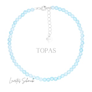 Könnte beinhalten: Eine zarte Halskette mit hellblauen, facettierten Perlen. Sie hat einen silbernen Verschluss und eine verstellbare Kette. Das Wort "TOPAS" wird in der Mitte angezeigt, mit dem Markennamen "Lauritas Schmuck" unten.