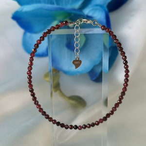 Könnte beinhalten: Ein zartes Armband aus roten Granatsteinen mit einem silbernen Herzanhänger und einem Karabinerverschluss.