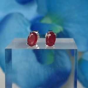 Op de afbeelding: Zilveren oorstekers met ovale robijn edelstenen. De rode edelstenen worden getoond op een helder acrylblok tegen een vage blauwe bloemenachtergrond. Een klassiek sieraad.