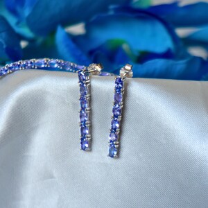 Op de afbeelding: Een set zilveren oorbellen en een armband met een rij lichtblauwe edelstenen. De sieraden worden getoond op een lichtblauwe satijnen stof.
