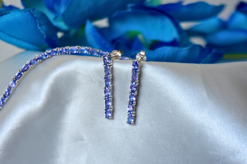 Op de afbeelding: Een set zilveren oorbellen en een armband met een rij lichtblauwe edelstenen. De sieraden worden getoond op een lichtblauwe satijnen stof.