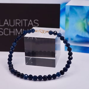 May include: A dark blue gemstone bracelet with a gold clasp. The bracelet is made of faceted beads and is displayed on a white surface.