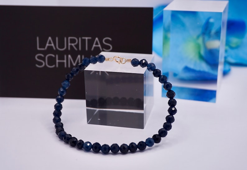 May include: A dark blue gemstone bracelet with a gold clasp. The bracelet is made of faceted beads and is displayed on a white surface.