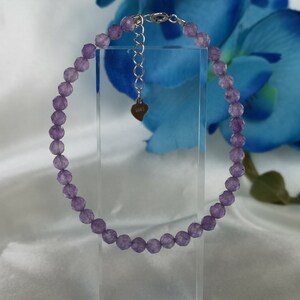 Könnte beinhalten: Ein zartes Armband aus lavendelfarbenen Perlen mit einer verstellbaren Kette in Silberoptik und einem herzförmigen Anhänger. Das Armband wird vor einem weißen Hintergrund mit einer blauen Blume präsentiert.