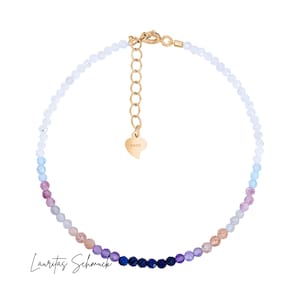 Könnte beinhalten: Ein zartes Perlenarmband mit einem Farbverlauf aus bunten Edelsteinen, der von Weiß zu Blautönen, Rosa und Lila übergeht. Das Armband hat einen goldfarbenen Verschluss und einen herzförmigen Anhänger. Der Text "Lauritas Schmuck" ist sichtbar.