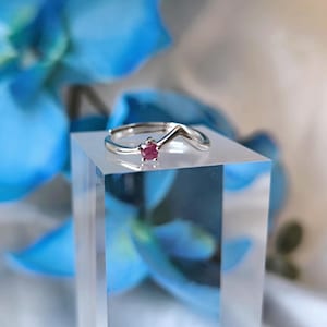 Op de afbeelding: Een zilveren ring met een kleine, rode edelsteen. De ring wordt getoond op een helder, vierkant blok. Het ontwerp is golvend. De achtergrond heeft vage blauwe bloemen en witte stof.