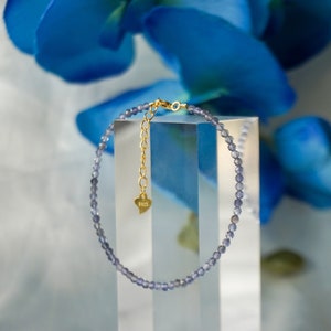 May include: A delicate bracelet made of small, light blue gemstone beads. The bracelet has a gold chain with a small heart charm that says "Bess".