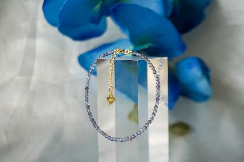 May include: A delicate bracelet made of small, light blue gemstone beads. The bracelet has a gold chain with a small heart charm that says "Bess".