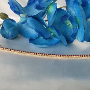 May include: A gold bracelet with a row of small, red gemstones. The bracelet is lying on a white surface.