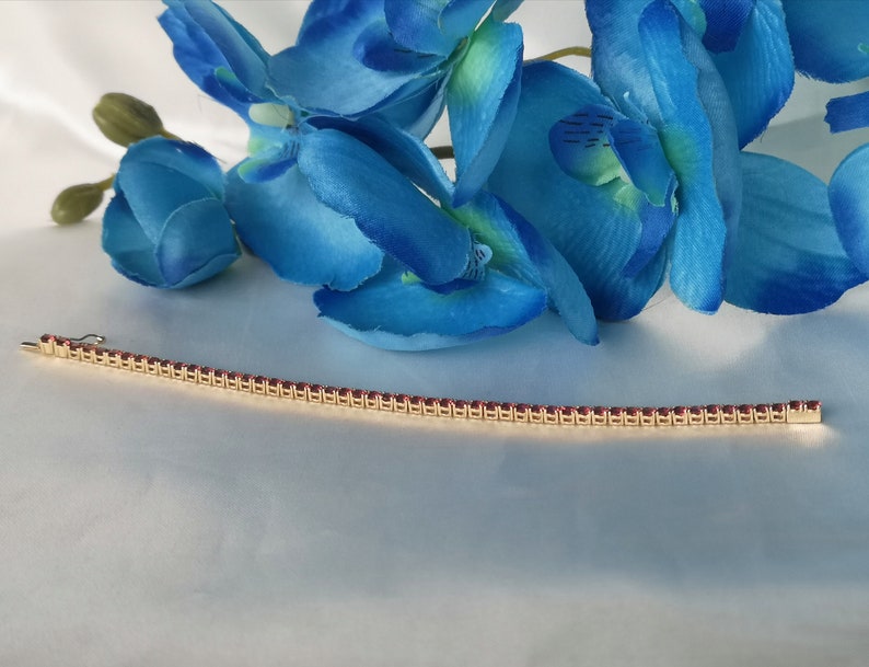 May include: A gold bracelet with a row of small, red gemstones. The bracelet is lying on a white surface.