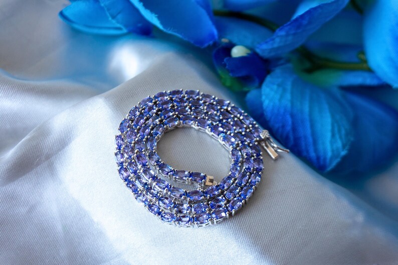 Op de afbeelding: Een zilveren ketting met een rij ovale, paarse edelstenen. De ketting ligt op een witte satijnen stoffen achtergrond.