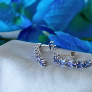 Op de afbeelding: Een paar zilveren creolen met een rij ovale blauwe edelstenen. De oorbellen liggen op een witte stoffen achtergrond.