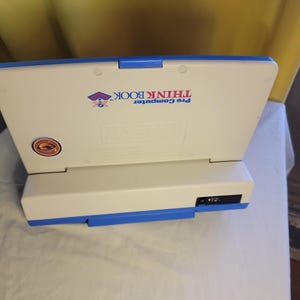 May include: A white and blue Pre Computer Think Book toy. The toy has a blue hinge and accents. The text "Pre Computer Think Book" is printed on the top. A sticker is on the lower left corner.