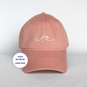 Può includere: Un berretto da baseball rosa con un design a onda ricamato in bianco. Il berretto ha una visiera curva e una cinghia regolabile.