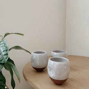 May include: Three handmade ceramic cups with a white glaze and a brown base are arranged on a wooden table. The cups have a rounded shape and a smooth, textured surface. The background is a neutral color.