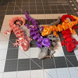 May include: Five 3D printed gecko lizards in different colors: pink, purple, orange, red, and gray. The geckos are posed in a variety of positions, with their tails curled up and their legs outstretched.