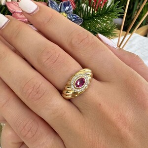 May include: A gold ring with a large oval ruby center stone and a halo of small diamonds. The ring has a ribbed design on the band and is set on a hand with white nail polish.