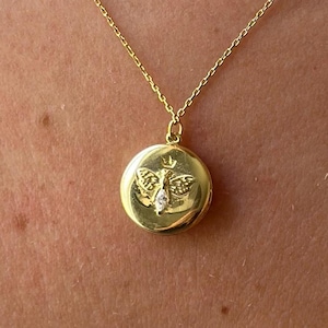 May include: Gold-coloured pendant necklace. The round pendant features a butterfly design with a small, clear gemstone in the centre. The necklace chain is delicate and also gold-coloured.