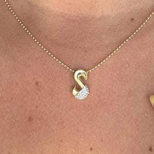 Puede incluir: Un collar dorado con un colgante en forma de la letra "S". El colgante está adornado con pequeñas piedras blancas brillantes. El collar se lleva alrededor del cuello de una persona.