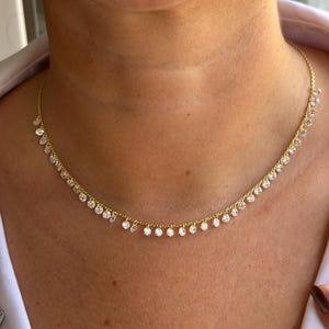 Puede incluir: Un delicado collar de oro con una serie de pequeñas piedras preciosas redondas y transparentes colgando de la cadena. El collar se lleva alrededor del cuello y las piedras preciosas captan la luz, creando un efecto brillante. El collar es una joya.