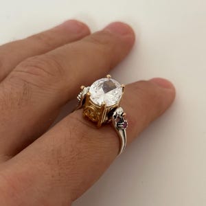 Könnte beinhalten: Ein silberner Ring mit einem großen, klaren ovalen Edelstein, der in einer goldenen Fassung sitzt. Der Ring hat zwei kleine rosafarbene Edelsteine auf der Ringschiene.
