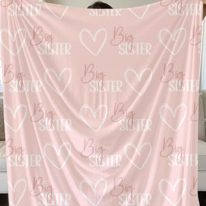May include: Pink fleece blanket with a repeating pattern of white hearts and the text "Big Sister" in a pink script font.
