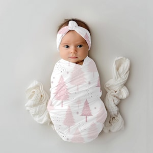 May include: A pink and white swaddle blanket with a pink Christmas tree pattern. A baby is wrapped in the swaddle and wearing a pink headband with a matching Christmas tree pattern.