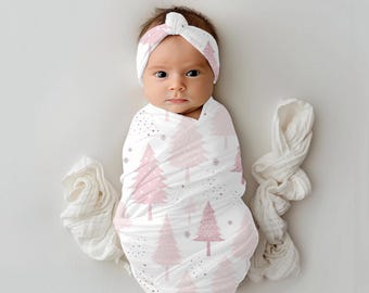 May include: A pink and white swaddle blanket with a pink Christmas tree pattern. A baby is wrapped in the swaddle and wearing a pink headband with a matching Christmas tree pattern.