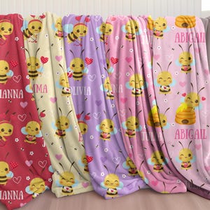 Pode incluir: Várias mantas de lã polar em tons de vermelho, creme, roxo e rosa, adornadas com um padrão repetido de abelhas de desenho animado sorridentes, corações e flores. Cada manta é personalizada com nomes como "IANNA", "EMMA", "OLIVIA", "INA", "TULA" e "ABIGAIL".