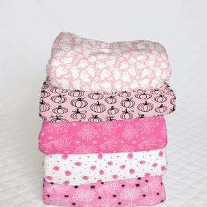 May include: A stack of five pink fabric squares with different Halloween-themed patterns. The patterns include ghosts, pumpkins, spiderwebs, and candy corn.