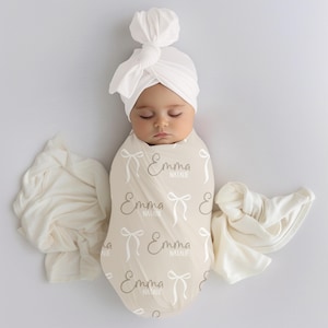 Könnte beinhalten: Ein schlafendes Baby, eingewickelt in eine beige Wickeldecke und mit einem weißen Turban-Hut. Die Decke ist mit weißen Schleifen und dem Namen "Emma Natalie" gemustert. Zusätzlicher cremefarbener Stoff ist um das Baby drapiert.