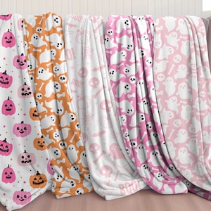 K&ouml;nnte beinhalten: Vier Halloween-Fleece-Decken mit verschiedenen Mustern. Eine Decke zeigt rosa K&uuml;rbisse, eine andere wei&szlig;e Geister, eine weitere rosa Sch&auml;del und Knochen und die letzte rosa Geister und Knochen.