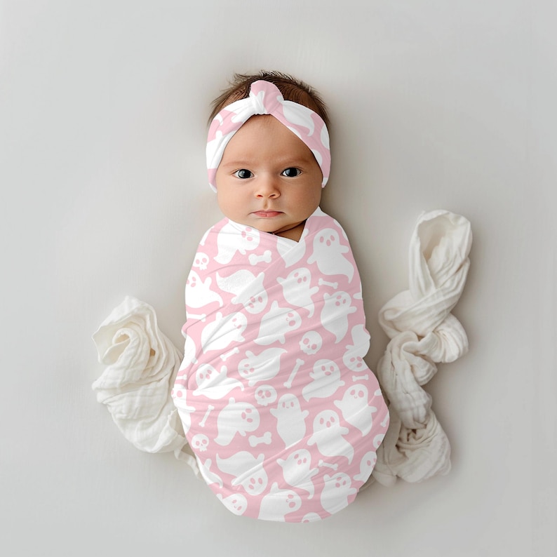 K&ouml;nnte beinhalten: Eine rosa Swaddle-Decke mit einem wei&szlig;en Geister- und Knochenmuster. Ein Baby tr&auml;gt die Swaddle und ein passendes rosa Stirnband mit einem wei&szlig;en Geistermuster.