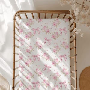 Puede incluir: Una sábana bajera blanca para cuna con un estampado repetido de rosas y lazos rosas. El diseño floral presenta racimos de rosas, intercalados con lazos rosas. La sábana está colocada dentro de una cuna de madera natural.