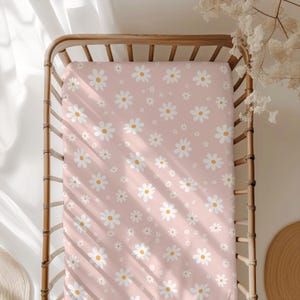 Peut inclure: Un drap-housse rose pour lit de bébé avec un motif de marguerites blanches. Le drap est fait d'un tissu doux et respirant, idéal pour une chambre de bébé.