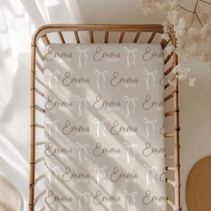 May include: A beige fitted crib sheet with a repeating pattern of white bows and the names "Emma Natalie" in brown script.