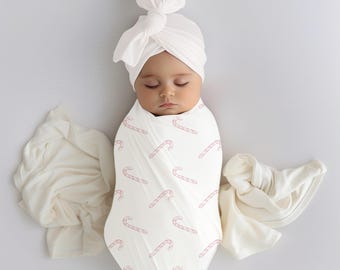 Könnte beinhalten: Ein schlafendes Baby, eingewickelt in eine weiße Wickeldecke mit rosa Zuckerstangenmuster, trägt einen passenden weißen Turbanhut. Das Baby ist von weichem, cremefarbenem Stoff umgeben.