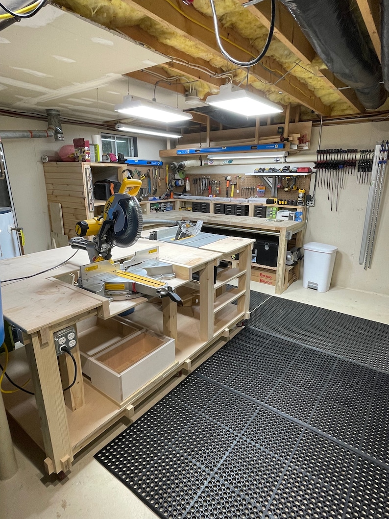 Puede incluir: Una sierra de inglete amarilla y negra montada en un banco de trabajo de madera con un caj&oacute;n. El banco de trabajo est&aacute; en un taller de s&oacute;tano con alfombras de goma negra. El taller tiene una variedad de herramientas y equipos en las paredes y estantes.