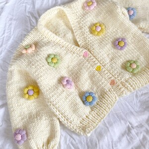 May include: A white knitted cardigan with a V-neck and colorful flower embellishments. The cardigan has a button closure and long sleeves.