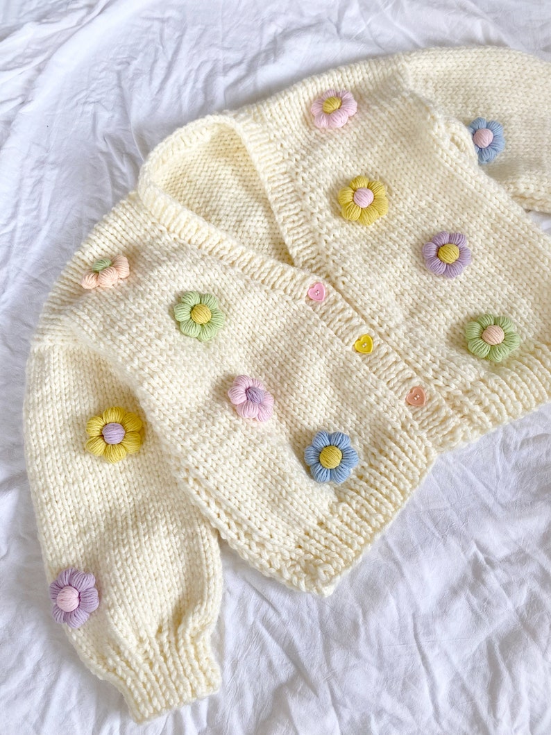 May include: A white knitted cardigan with a V-neck and colorful flower embellishments. The cardigan has a button closure and long sleeves.