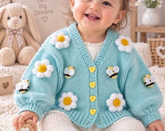 May include: A light blue knit cardigan featuring three-dimensional white daisy appliqués with yellow centers and small bumblebee appliqués. The garment has a V-neckline, ribbed cuffs and hem, and four yellow heart-shaped buttons down the front.