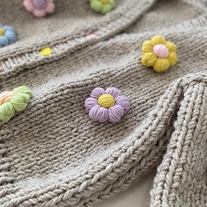 May include: A gray knitted cardigan with colorful crocheted flower embellishments. The cardigan has a button closure and a ribbed texture.