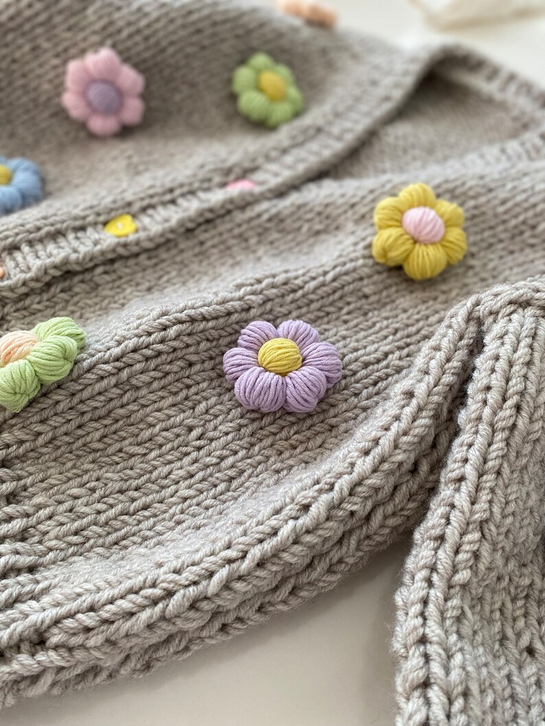 May include: A gray knitted cardigan with colorful crocheted flower embellishments. The cardigan has a button closure and a ribbed texture.