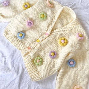 May include: A white knitted cardigan with a V-neck and long sleeves. The cardigan is decorated with colorful flower appliques.