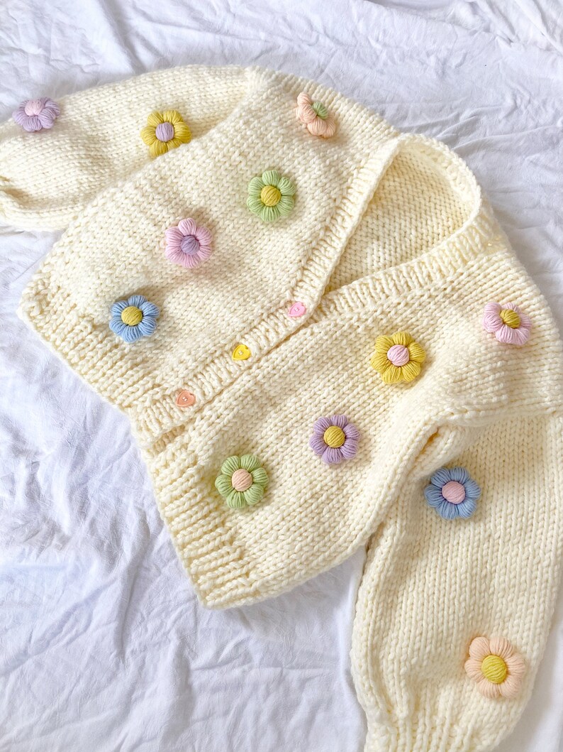 May include: A white knitted cardigan with a V-neck and long sleeves. The cardigan is decorated with colorful flower appliques.