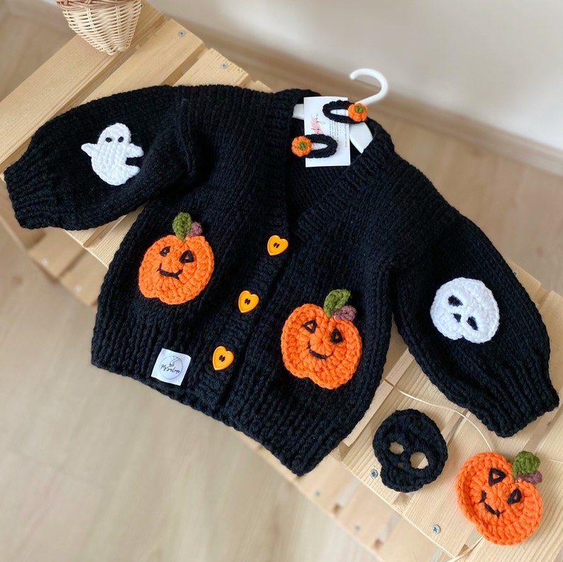 May include: Black knitted cardigan with pumpkin and ghost appliques. The cardigan has heart-shaped buttons and comes with a matching pumpkin and ghost hair clip set.