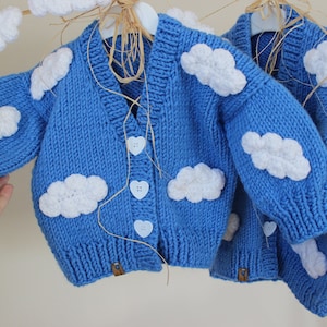 Puede incluir: Un cárdigan azul de punto con nubes blancas de ganchillo y botones en forma de corazón.