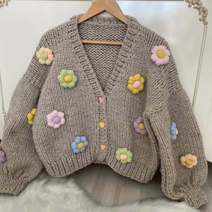 May include: A beige knitted cardigan with a V-neck and long sleeves. The cardigan is decorated with colorful flower appliques.